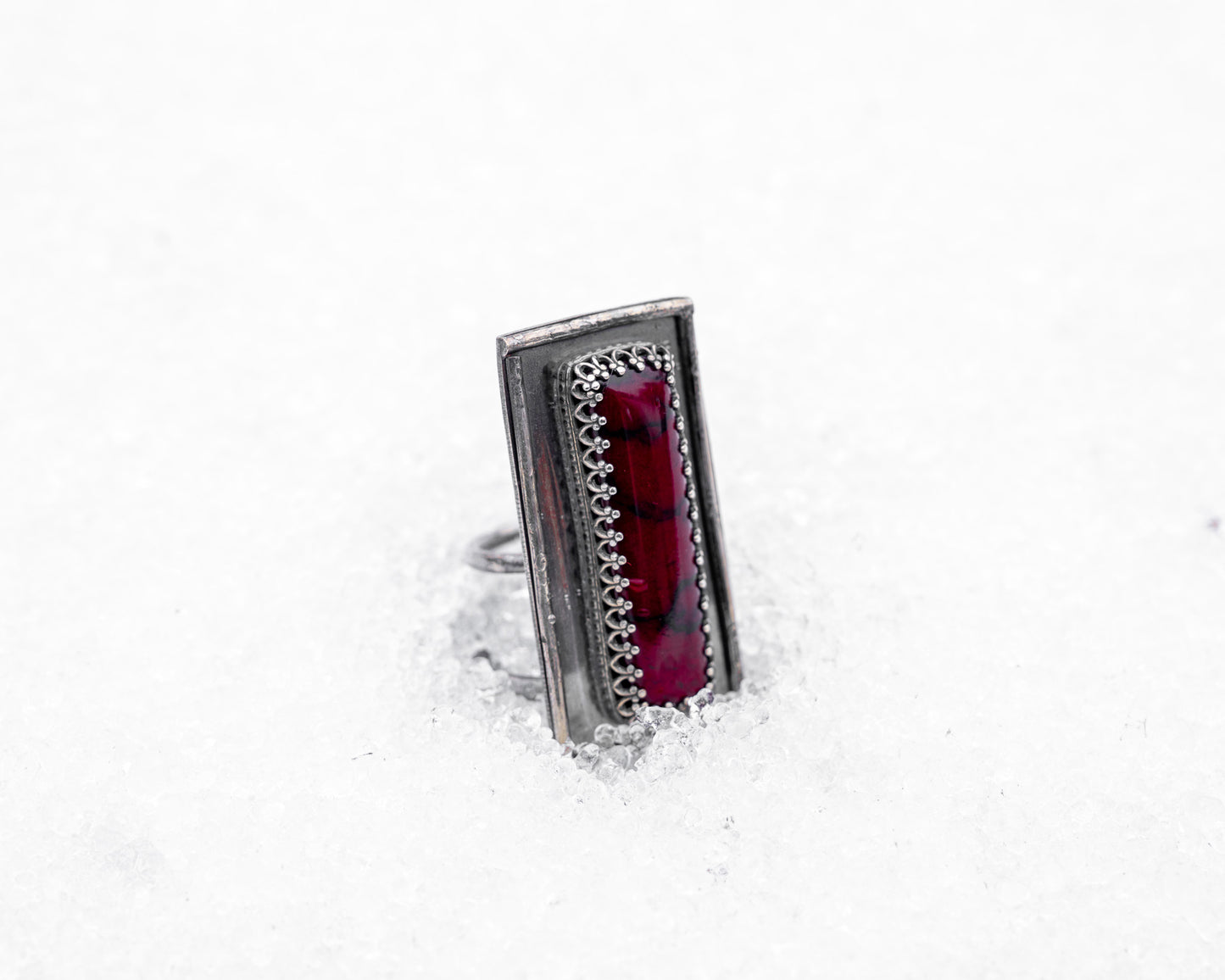 Close-up of a sterling silver ring with an elongated rectangular design set with a deep red bloody bison jasper, nestled upright in textured white snow. The ring has detailed wirework around the stone and a dark, oxidized finish, evoking an ancient, blood-bound artifact.