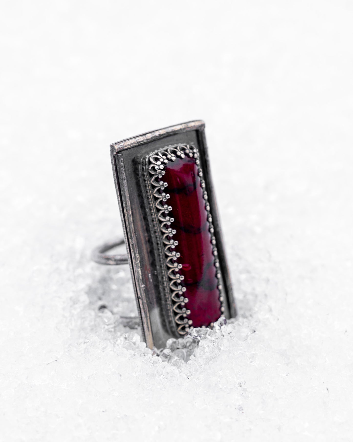 Close-up of a sterling silver ring with an elongated rectangular design set with a deep red bloody bison jasper, nestled upright in textured white snow. The ring has detailed wirework around the stone and a dark, oxidized finish, evoking an ancient, blood-bound artifact.