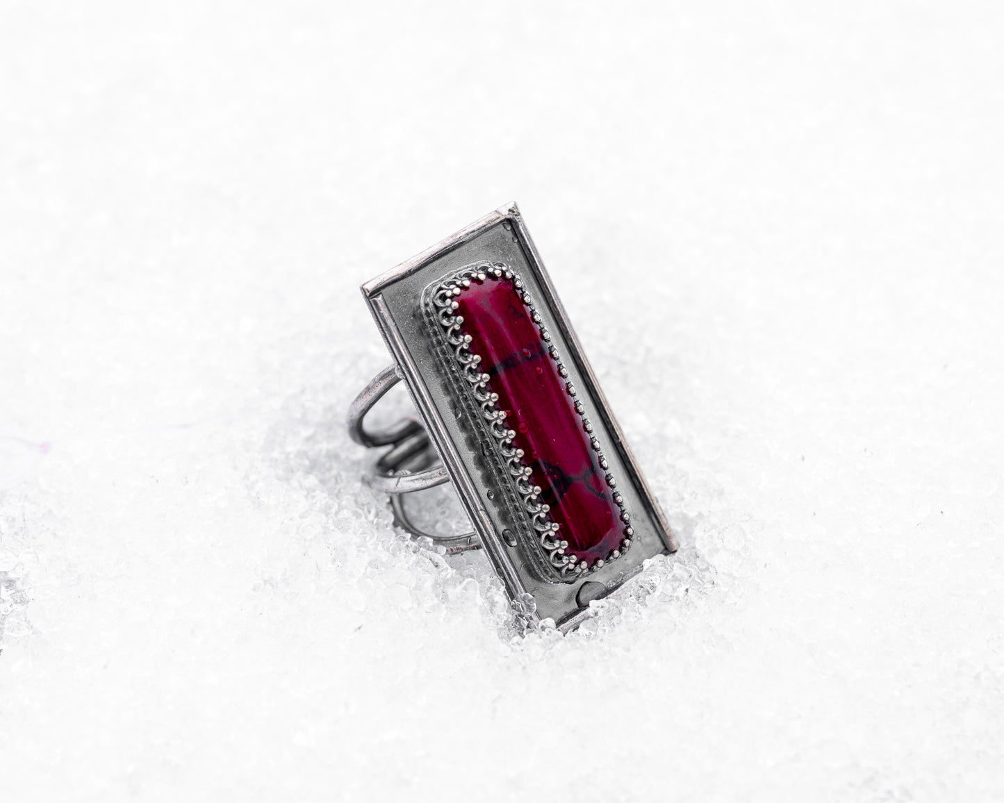 Close-up of a sterling silver ring with an elongated rectangular design set with a deep red bloody bison jasper, nestled upright in textured white snow. The ring has detailed wirework around the stone and a dark, oxidized finish, evoking an ancient, blood-bound artifact.