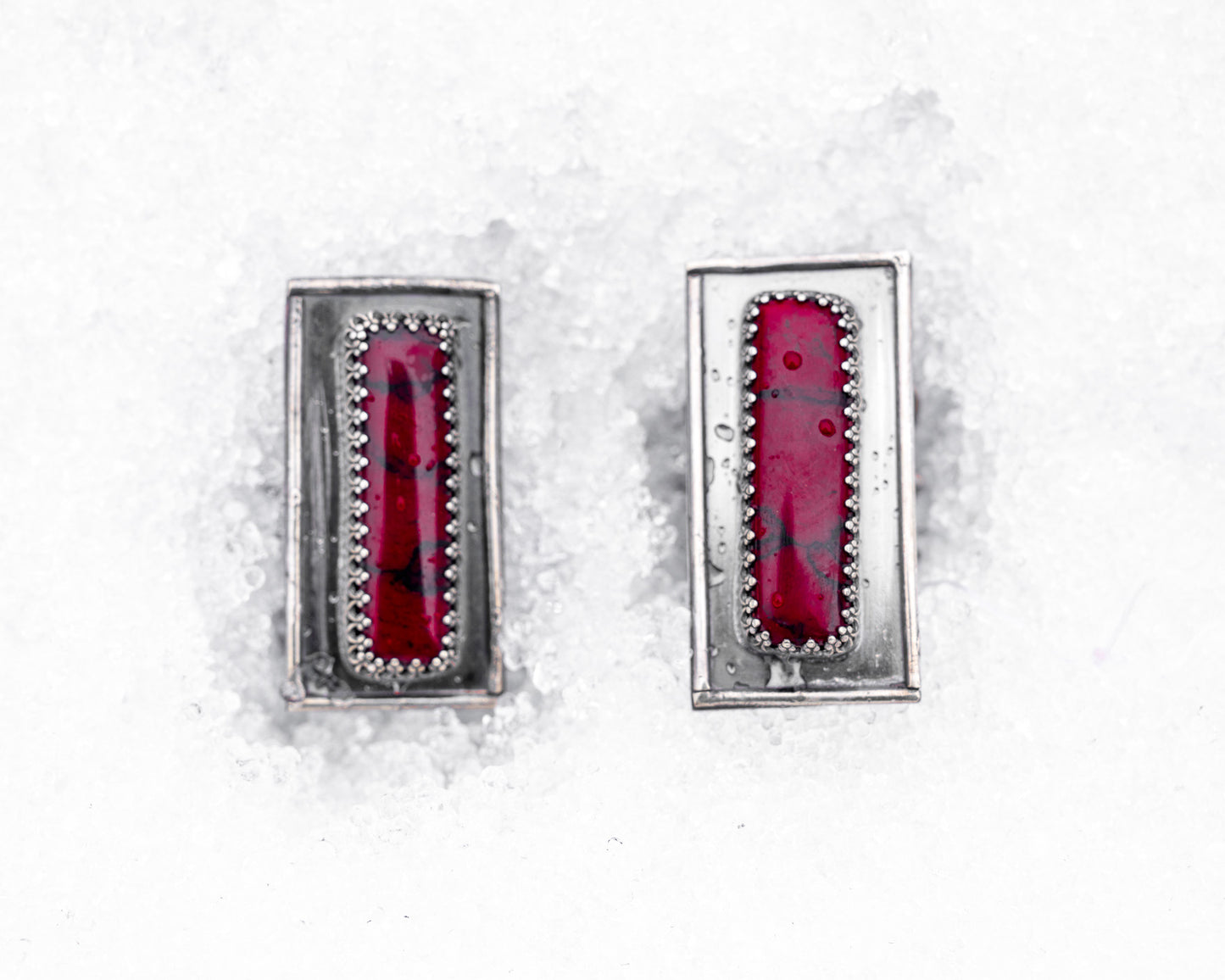 Close-up of a sterling silver ring with an elongated rectangular design set with a deep red bloody bison jasper, nestled upright in textured white snow. The ring has detailed wirework around the stone and a dark, oxidized finish, evoking an ancient, blood-bound artifact.