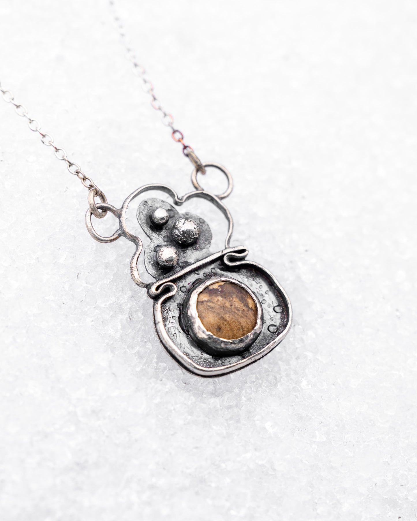 Handcrafted sterling silver necklace with a rounded pendant featuring three raised silver bubbles above a bezel-set garden jasper cabochon. Styled on snow, the pendant resembles a bubbling cauldron used in magical rituals.