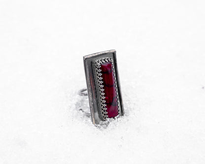 Close-up of a sterling silver ring with an elongated rectangular design set with a deep red bloody bison jasper, nestled upright in textured white snow. The ring has detailed wirework around the stone and a dark, oxidized finish, evoking an ancient, blood-bound artifact.
