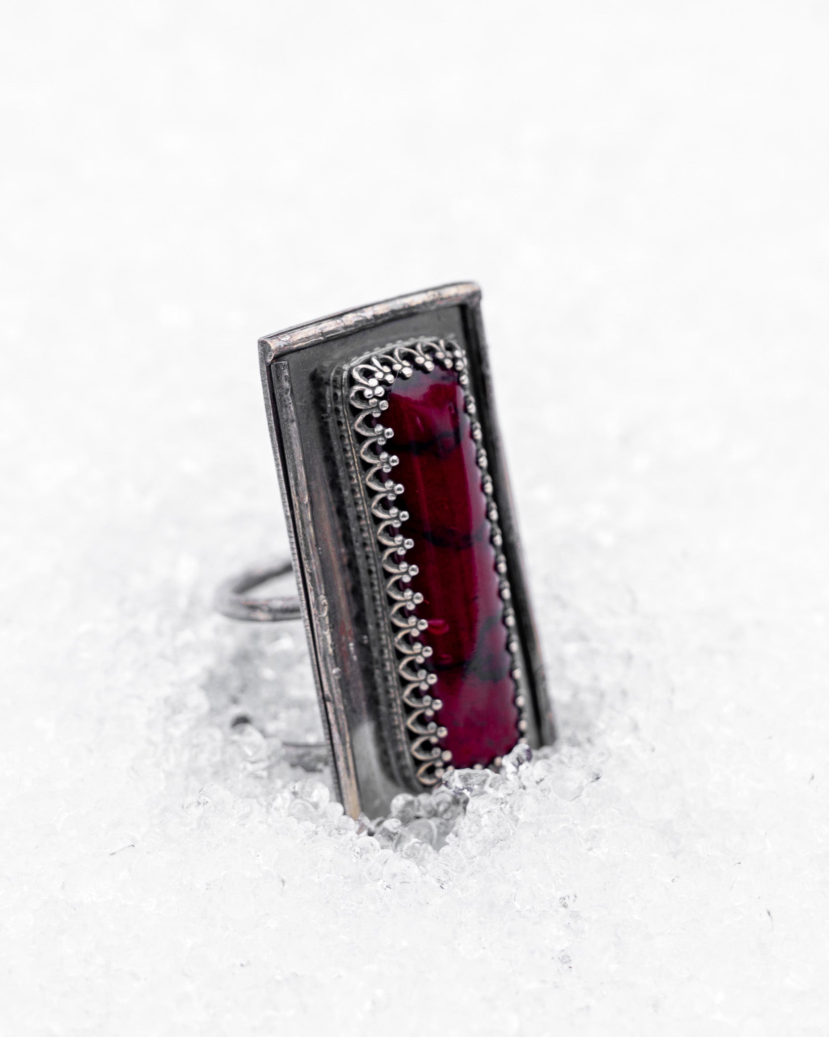 Close-up of a sterling silver ring with an elongated rectangular design set with a deep red bloody bison jasper, nestled upright in textured white snow. The ring has detailed wirework around the stone and a dark, oxidized finish, evoking an ancient, blood-bound artifact.
