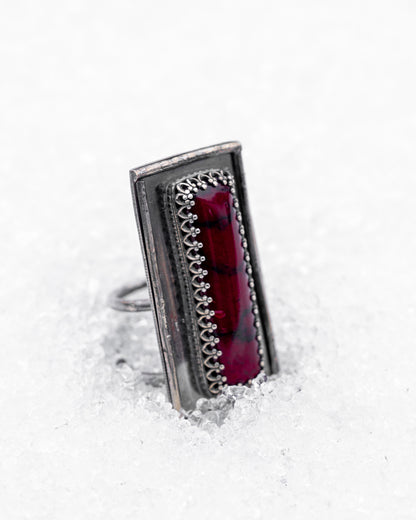 Close-up of a sterling silver ring with an elongated rectangular design set with a deep red bloody bison jasper, nestled upright in textured white snow. The ring has detailed wirework around the stone and a dark, oxidized finish, evoking an ancient, blood-bound artifact.
