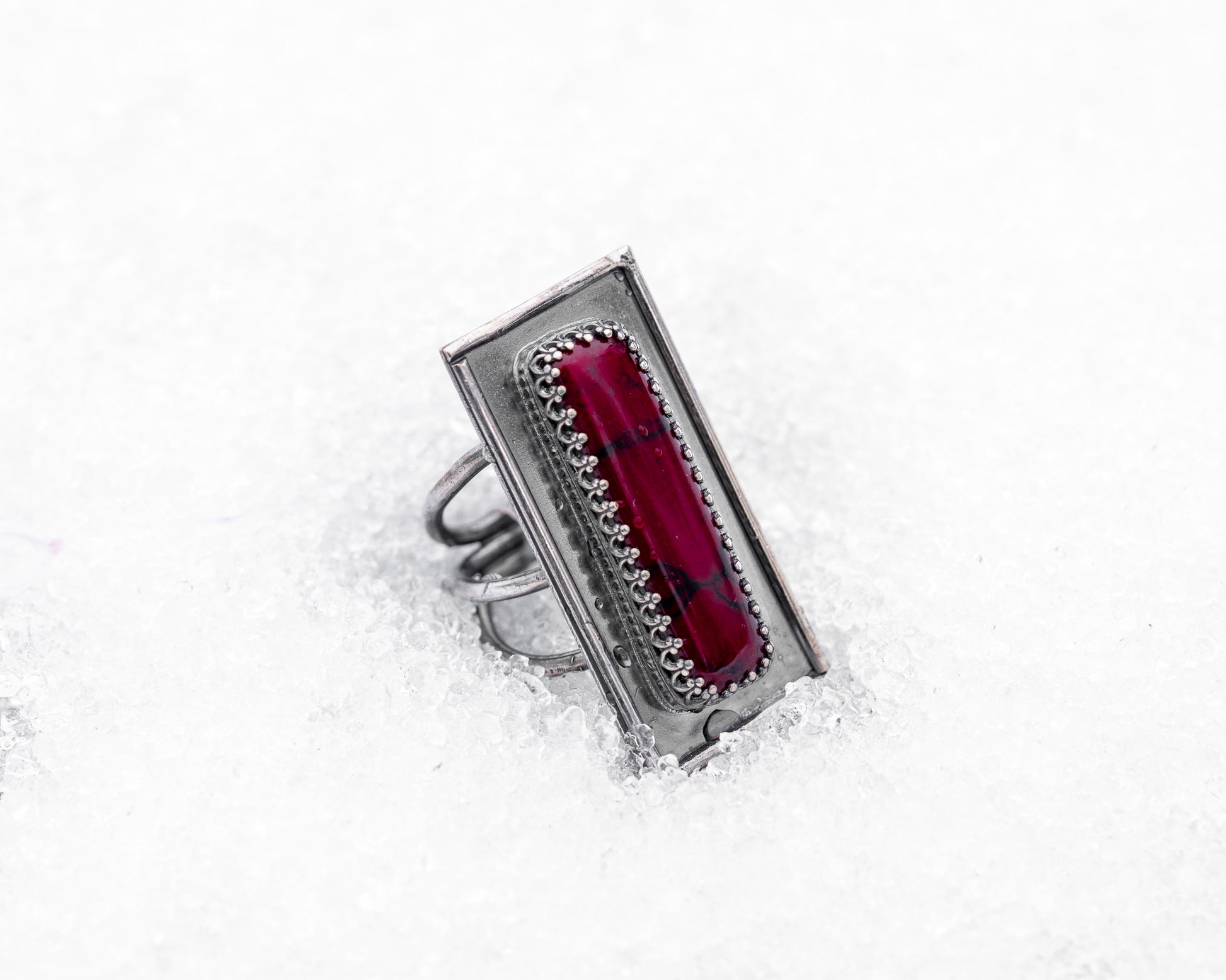 Close-up of a sterling silver ring with an elongated rectangular design set with a deep red bloody bison jasper, nestled upright in textured white snow. The ring has detailed wirework around the stone and a dark, oxidized finish, evoking an ancient, blood-bound artifact.
