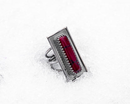 Close-up of a sterling silver ring with an elongated rectangular design set with a deep red bloody bison jasper, nestled upright in textured white snow. The ring has detailed wirework around the stone and a dark, oxidized finish, evoking an ancient, blood-bound artifact.
