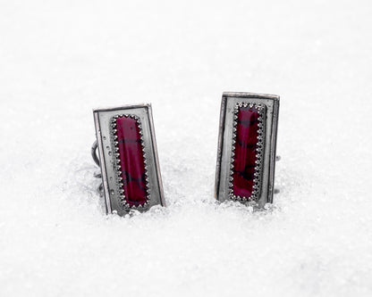Close-up of a sterling silver ring with an elongated rectangular design set with a deep red bloody bison jasper, nestled upright in textured white snow. The ring has detailed wirework around the stone and a dark, oxidized finish, evoking an ancient, blood-bound artifact.
