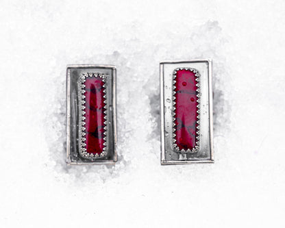 Close-up of a sterling silver ring with an elongated rectangular design set with a deep red bloody bison jasper, nestled upright in textured white snow. The ring has detailed wirework around the stone and a dark, oxidized finish, evoking an ancient, blood-bound artifact.
