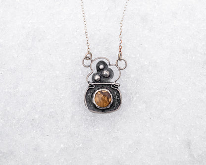 Handcrafted sterling silver necklace with a rounded pendant featuring three raised silver bubbles above a bezel-set garden jasper cabochon. Styled on snow, the pendant resembles a bubbling cauldron used in magical rituals.
