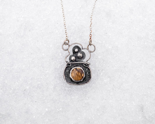 Handcrafted sterling silver necklace with a rounded pendant featuring three raised silver bubbles above a bezel-set garden jasper cabochon. Styled on snow, the pendant resembles a bubbling cauldron used in magical rituals.
